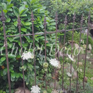 Dekoratives Rankgitter aus dunkelrostfarben patiniertem Eisen vor grüner Gartenbepflanzung.